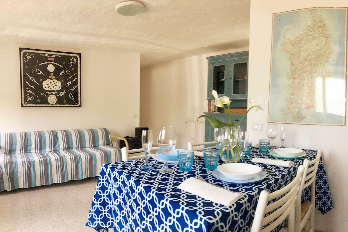 A dining area is presented with a blue-patterned tablecloth, set with plates and glasses, ready for a meal. A striped sofa adds seating, while a map of Sardinia decorates the wall, contributing to the home's coastal theme.