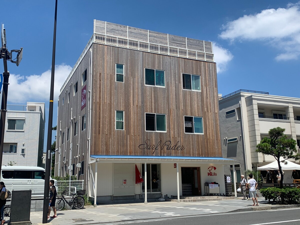 The accommodation features a three-story wooden building with a contemporary design. The facade showcases light brown wood, giving it a natural look. The entrance is marked by a sign, and outdoor seating is visible on the sidewalk, surrounded by greenery.