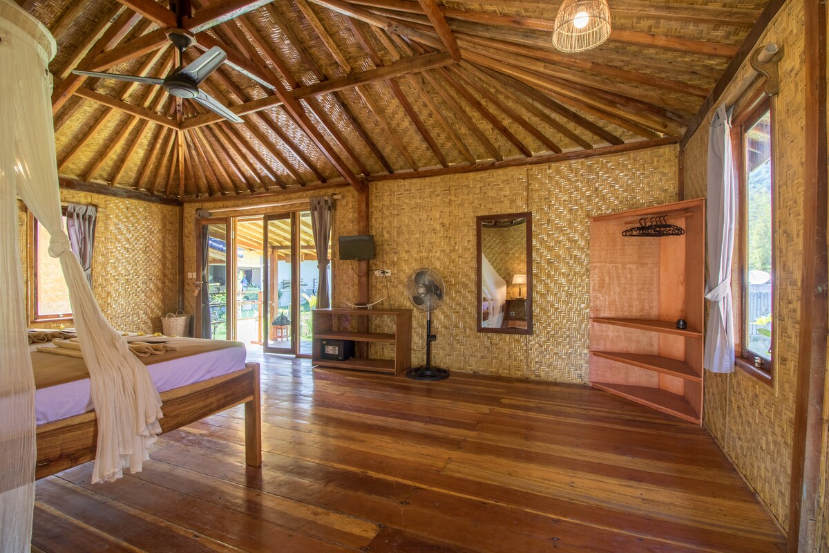 A spacious bungalow interior features woven bamboo walls and a high ceiling with wooden beams. A large double bed is positioned near glass doors that open to an outdoor area, allowing natural light to brighten the space. Additional furnishings include a wooden dresser and a ceiling fan.