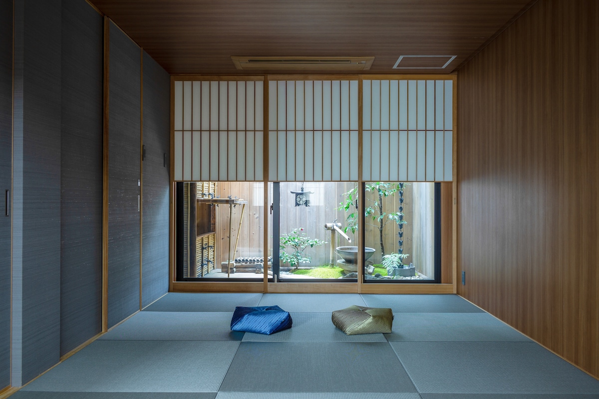 A serene room is highlighted by tatami flooring and wooden walls. Large sliding glass doors reveal a Japanese garden, while three decorative cushions in blue and gold are placed on the floor, inviting relaxation and contemplation.