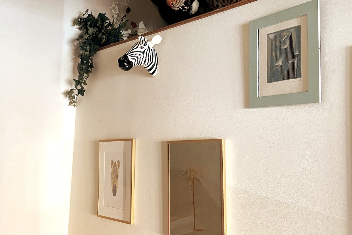 A wall features a variety of framed artwork, including illustrations and prints, arranged in an attractive layout. A decorative zebra head mounted above the frames adds a playful touch. Greenery is present, subtly enhancing the visual appeal of the space.