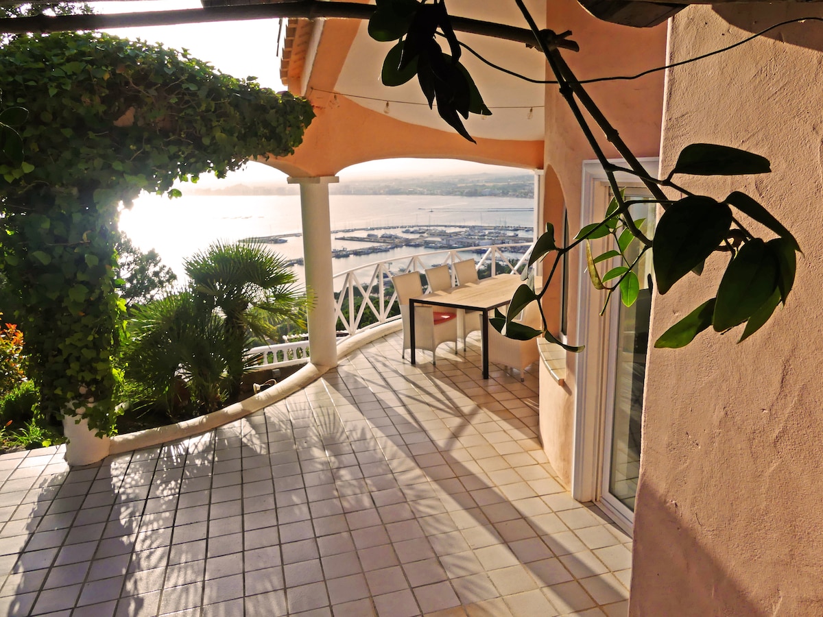 An outdoor terrace is visible, framed by greenery. A wooden table and chairs are positioned for dining, overlooking a scenic view of the marina and water. The space features tiled flooring and is bathed in natural light, enhancing the tranquil atmosphere.