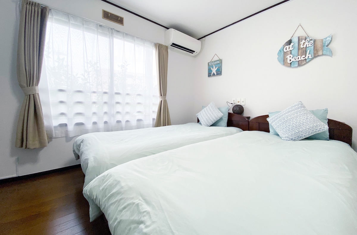 The room features two single beds with light-colored bedding and decorative pillows. Natural light filters through a sheer curtain, which enhances the bright and airy feel. A small wall decor piece reading 'at the Beach' adds a coastal touch.