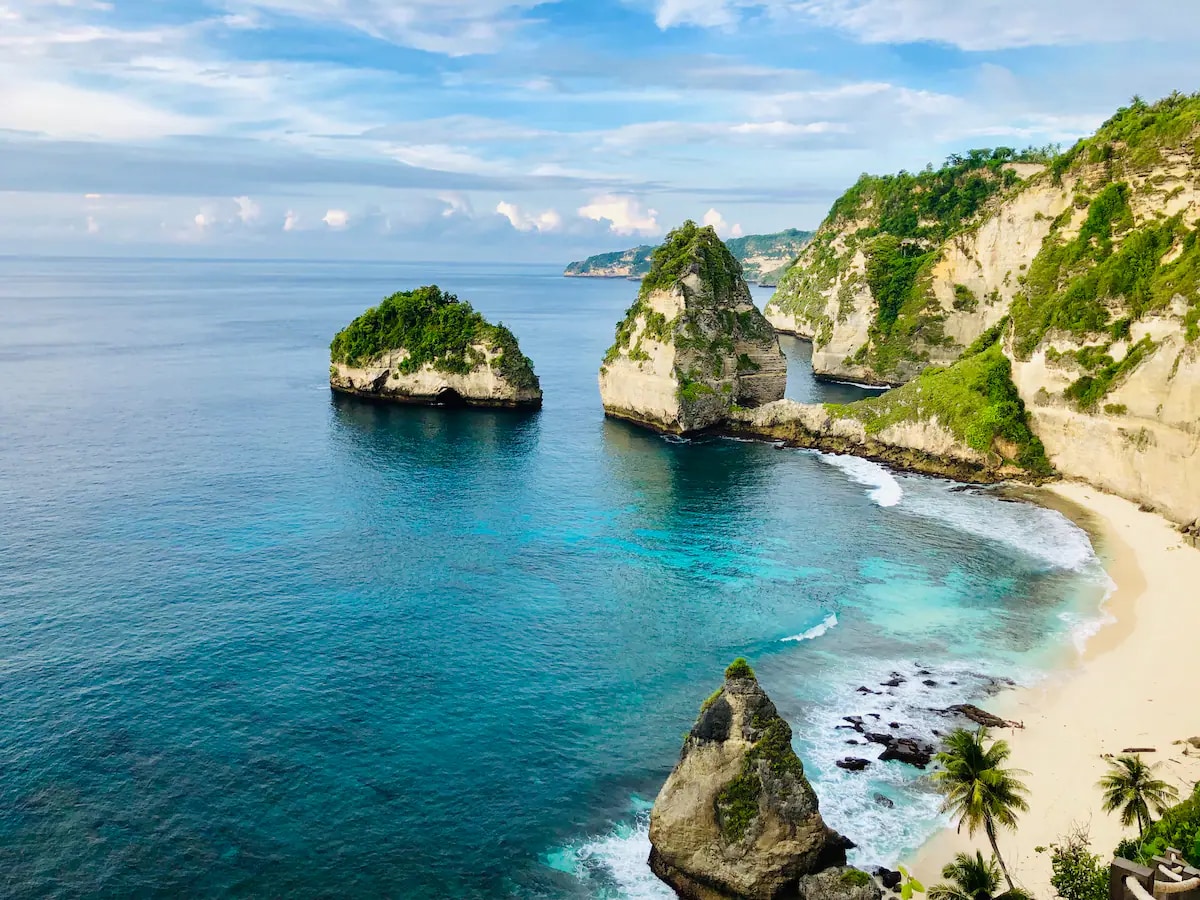 A serene coastal landscape is displayed, featuring crystal-clear turquoise waters lapping against sandy beaches. Towering cliffs covered in lush greenery rise majestically, and small rocky islands are seen in the distance. A gentle blend of blues and greens creates a tranquil scene.