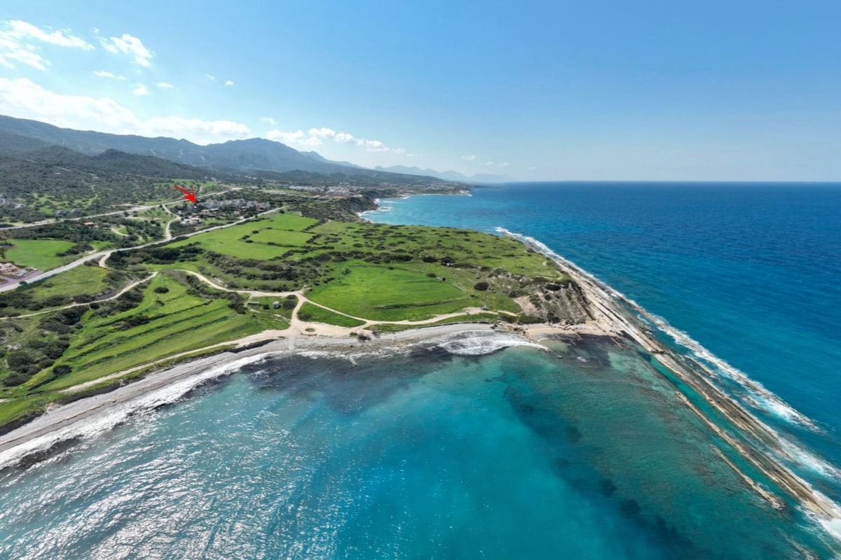 Aerial view showcasing the stunning coastline, clear turquoise waters, and lush green land of Akanthou. The beach meets rocky formations under the bright blue sky, with mountains visible in the background, enhancing the natural beauty of the area.