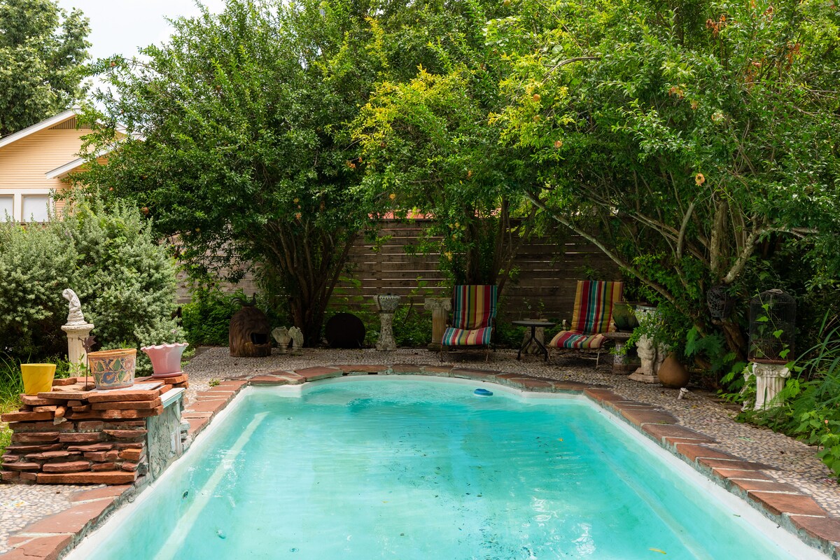 A serene swimming pool is framed by lush greenery and vibrant plants. Two colorful lounge chairs are positioned nearby under the shade of trees, providing a relaxing spot for guests. The pool area is designed with natural stone details, creating a welcoming outdoor space.