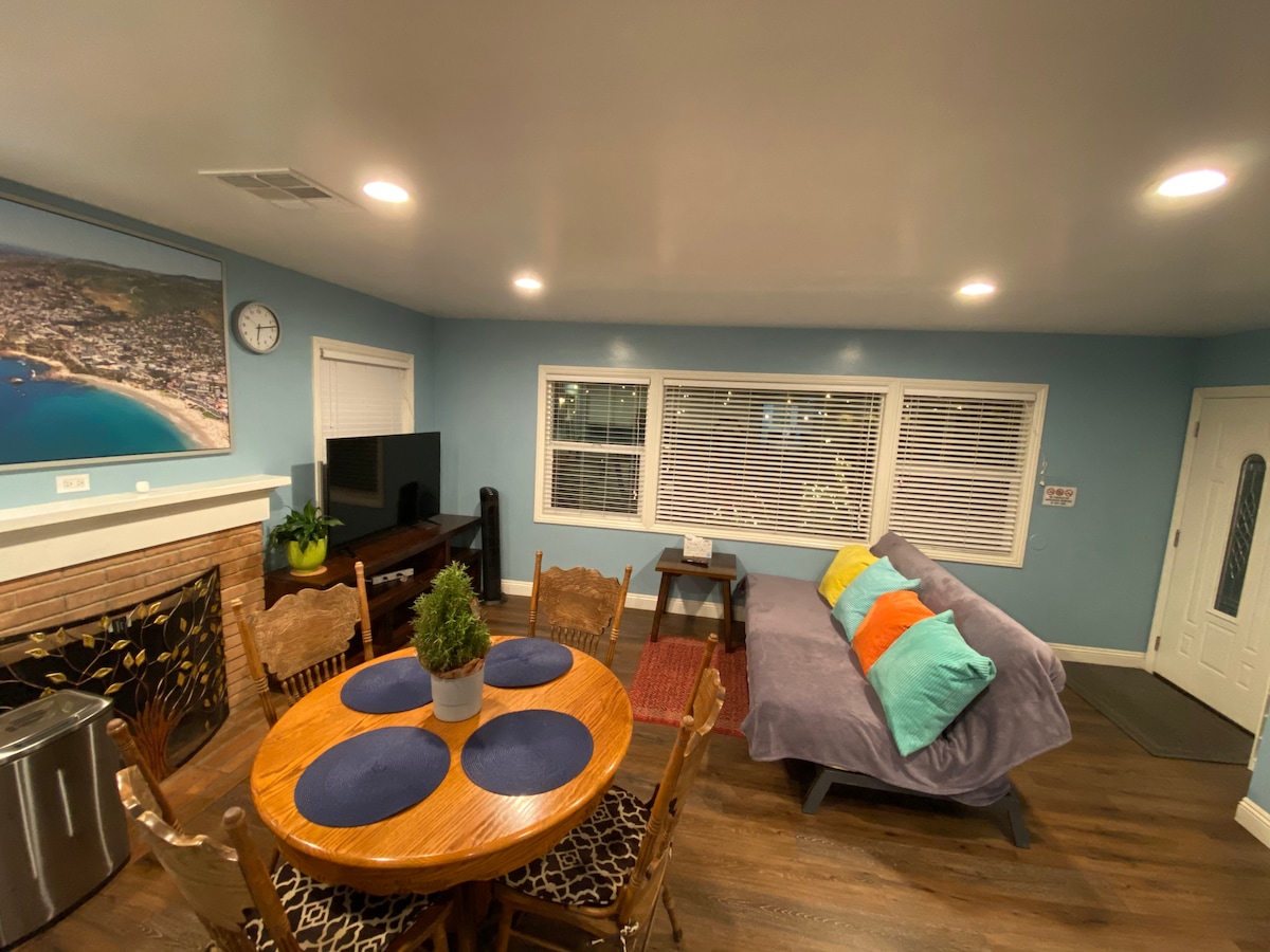 A welcoming living area is presented, featuring a round wooden dining table with blue placemats and four wooden chairs. A comfortable couch with bright cushions is situated alongside a 49" TV mounted on the wall, and large windows provide natural light.