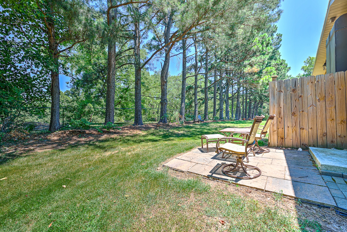 A back patio area is equipped with two comfortable lounge chairs, positioned on a stone surface. The outdoor space is surrounded by lush greenery and tall trees, providing a peaceful, natural setting. A wooden fence encloses the area for added privacy.