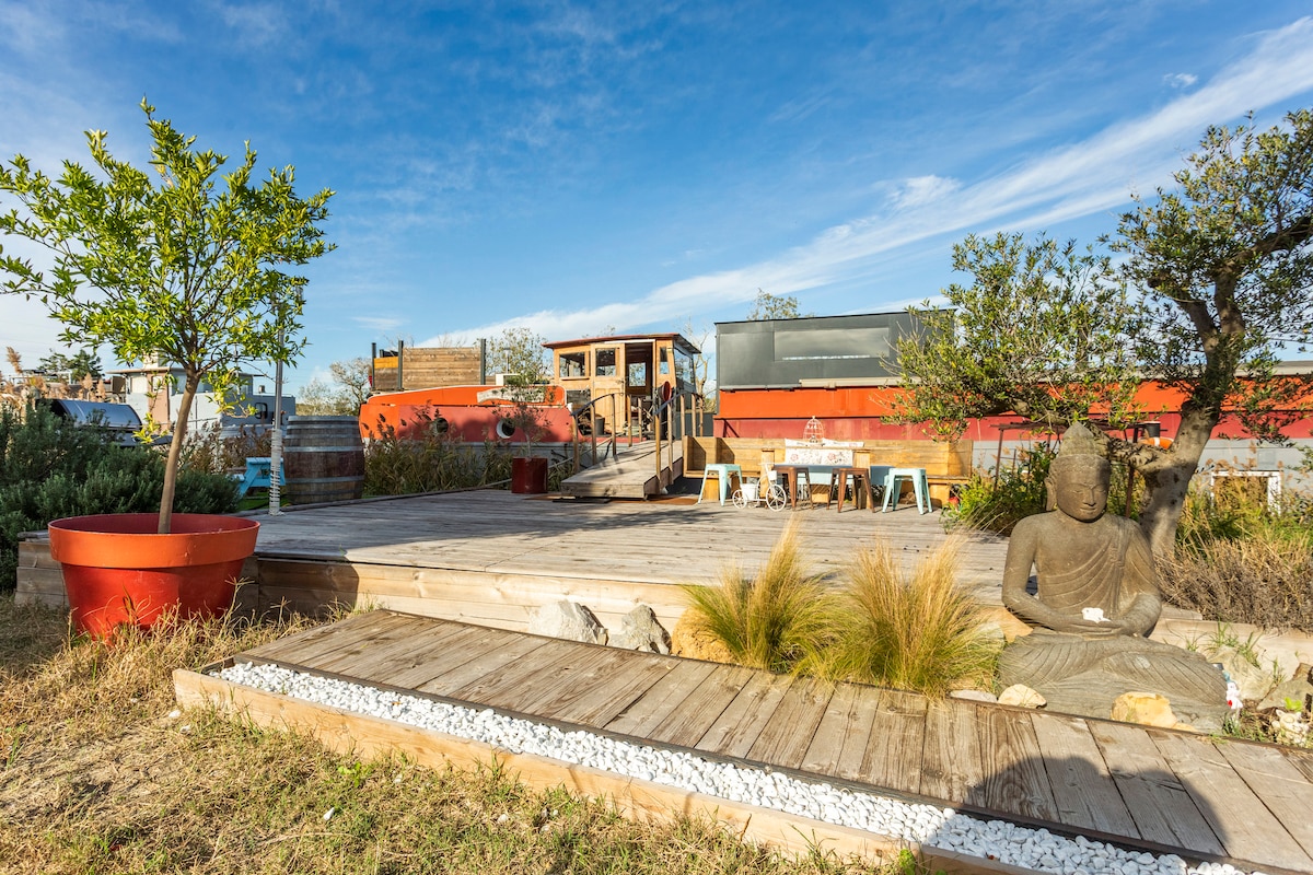 A wooden deck area is surrounded by lush greenery, featuring a large planter and a decorative statue. A dining table and chairs are positioned against the backdrop of a vintage ship, offering a serene outdoor space for relaxation.