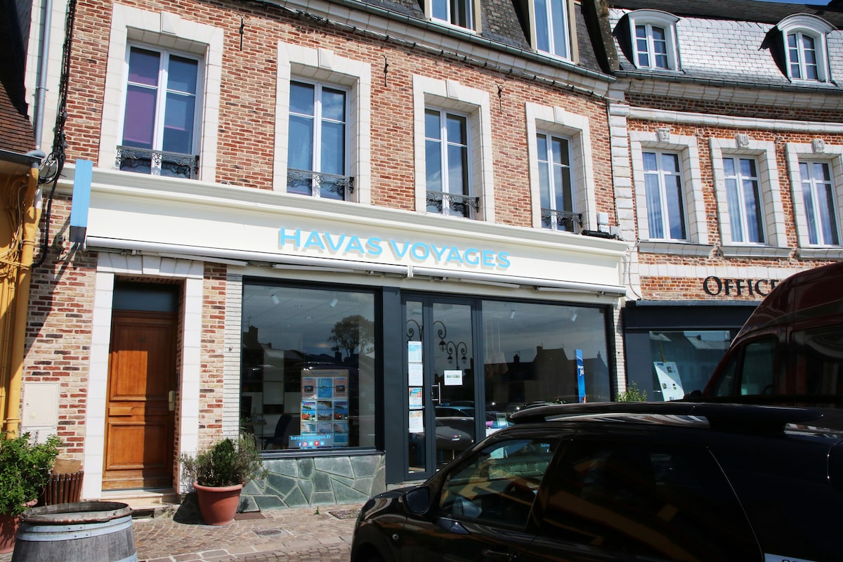 A brick facade features large windows in a commercial space, displaying travel brochures and a welcoming entrance. The shop's name, 'Havas Voyages,' is prominently mounted above the entrance. Nearby, a parked car is visible, and planters add greenery to the scene.