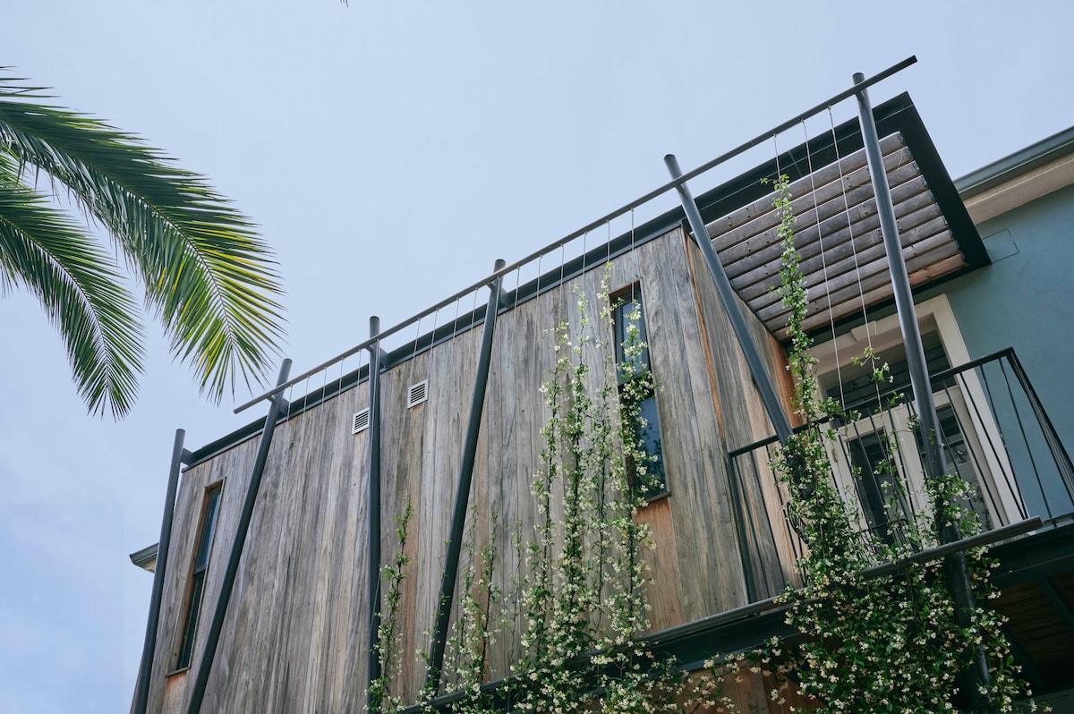 The exterior of Harpers House showcases a contemporary design that harmonizes with its surroundings. Wooden panels and greenery adorn the facade, while balconies and windows reflect elevated architectural features. The image captures the interplay of natural elements and modern aesthetics.