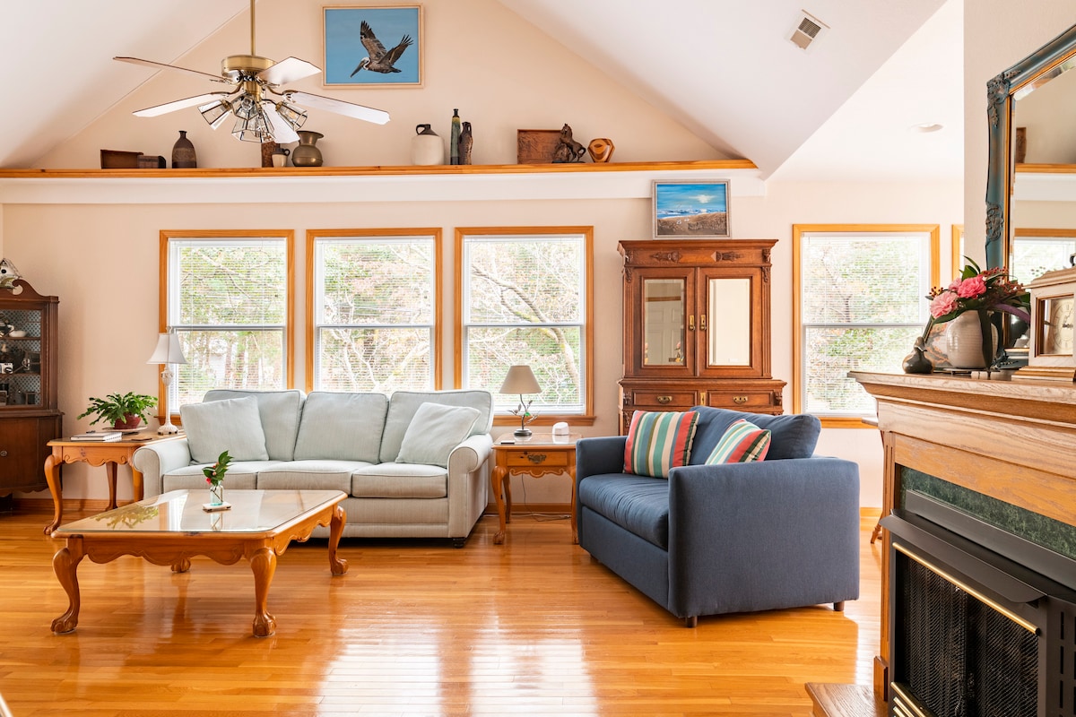 The great room is filled with natural light, featuring large windows overlooking a wooded area. A light-colored sofa and a dark blue love seat are positioned around a wooden coffee table. A decorative cabinet and an ornate fireplace add character to the space.