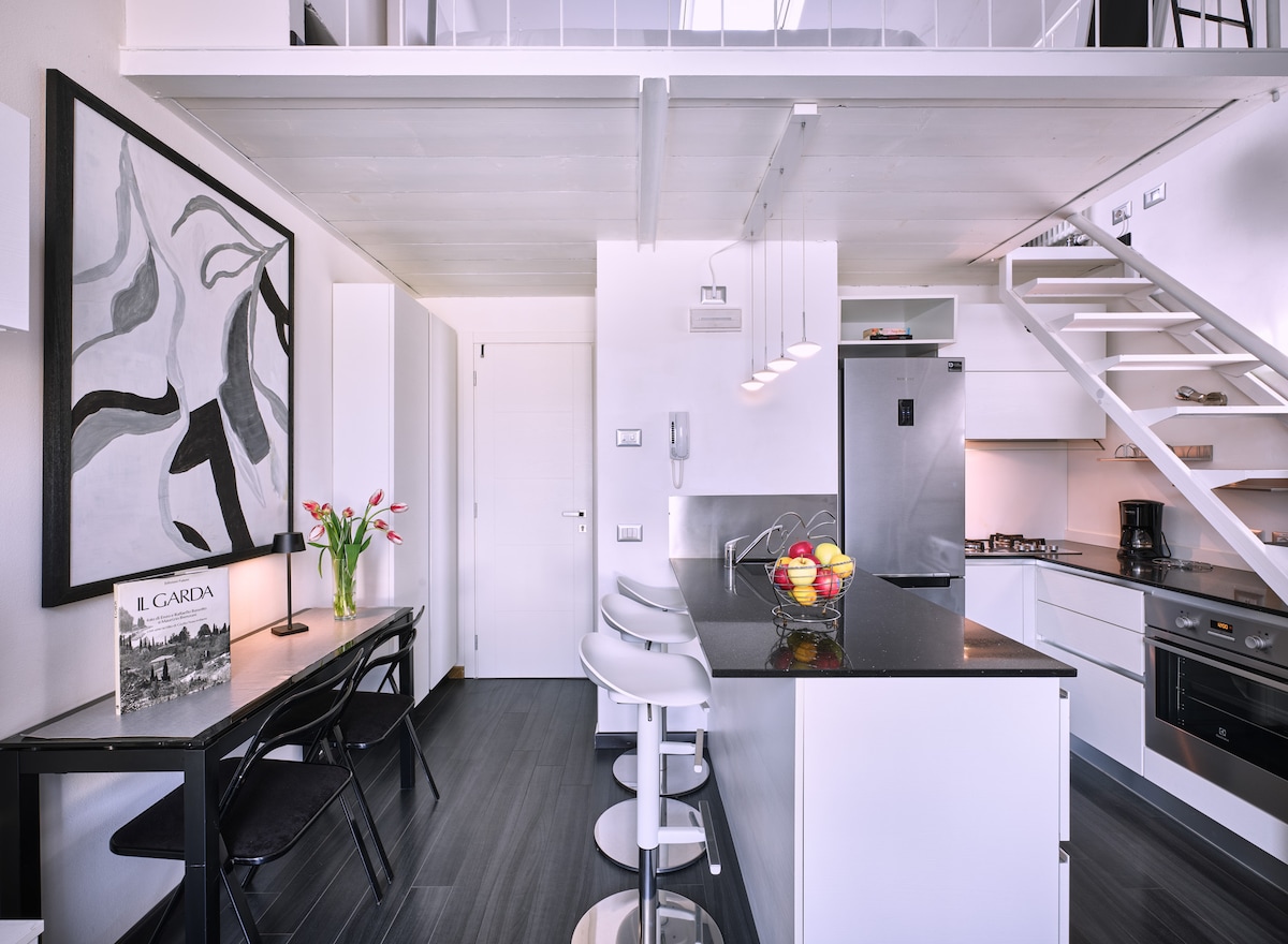 The modern kitchen area features sleek black countertops with a stylish bar and three high stools. A dining table with black chairs sits beside a wall adorned with abstract artwork. A staircase leads to the bright mezzanine where the sleeping area is located.