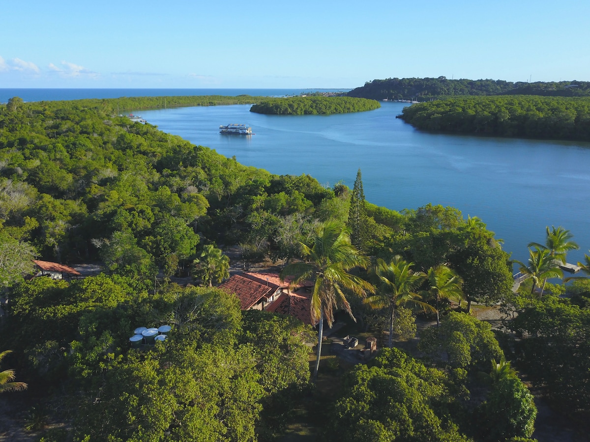 An aerial view showcases the lush greenery surrounding the calm waters of the João de Tiba river. The landscape features islands and a distant boat, offering a serene perspective of nature's beauty interspersed with palm trees and dense vegetation.
