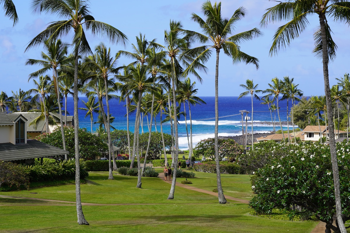 Lush greenery surrounds a pathway that meanders through palm trees, leading towards the vibrant blue Pacific Ocean. Gentle waves can be seen crashing along the shoreline, with a clear sky above enhancing the coastal view.