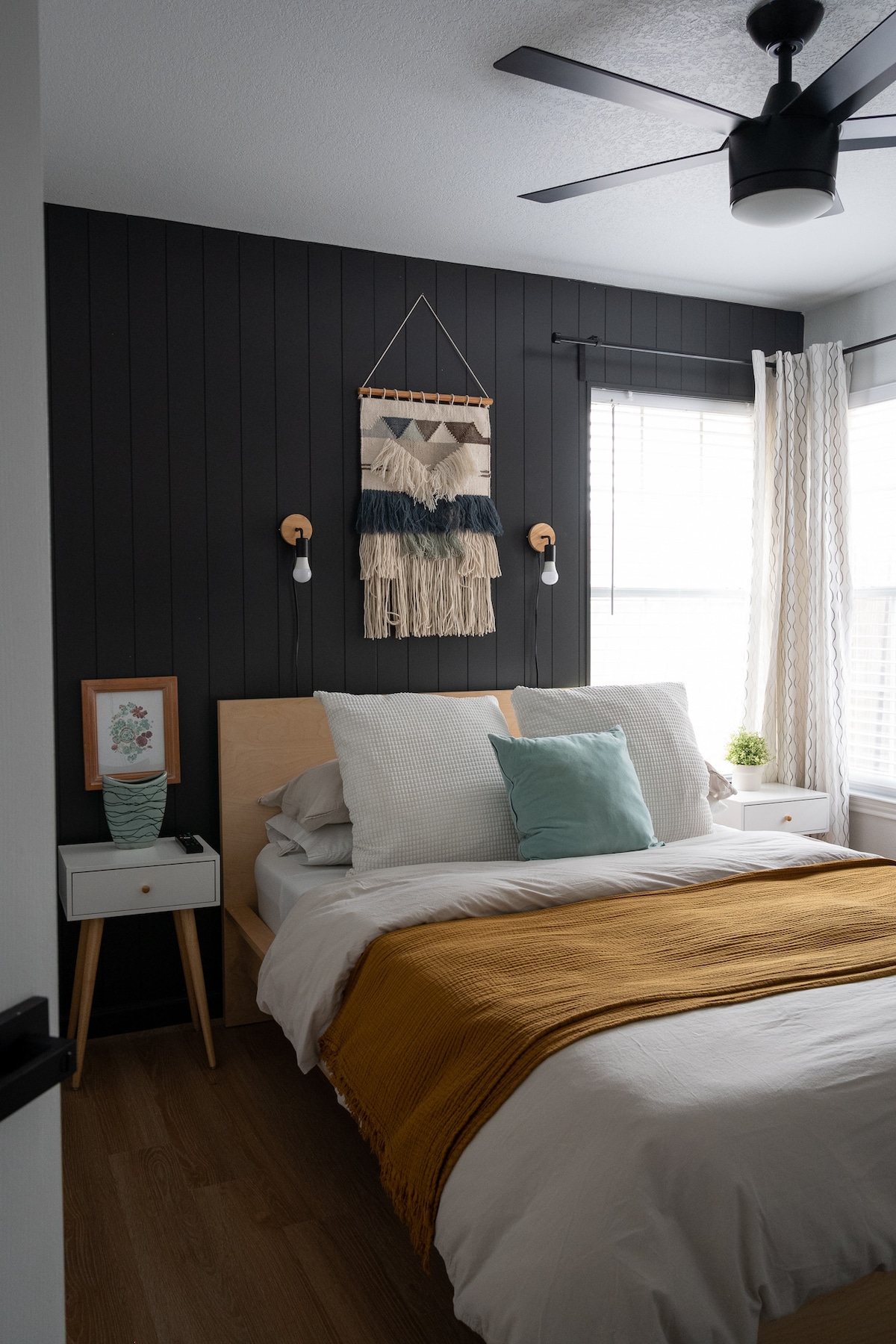 A cozy bedroom is featured with a spacious bed dressed in white linens and a mustard-colored throw. A macrame wall hanging adds texture to the dark accent wall. Natural light filters through the windows, illuminating the minimalist nightstand and decorative accents.