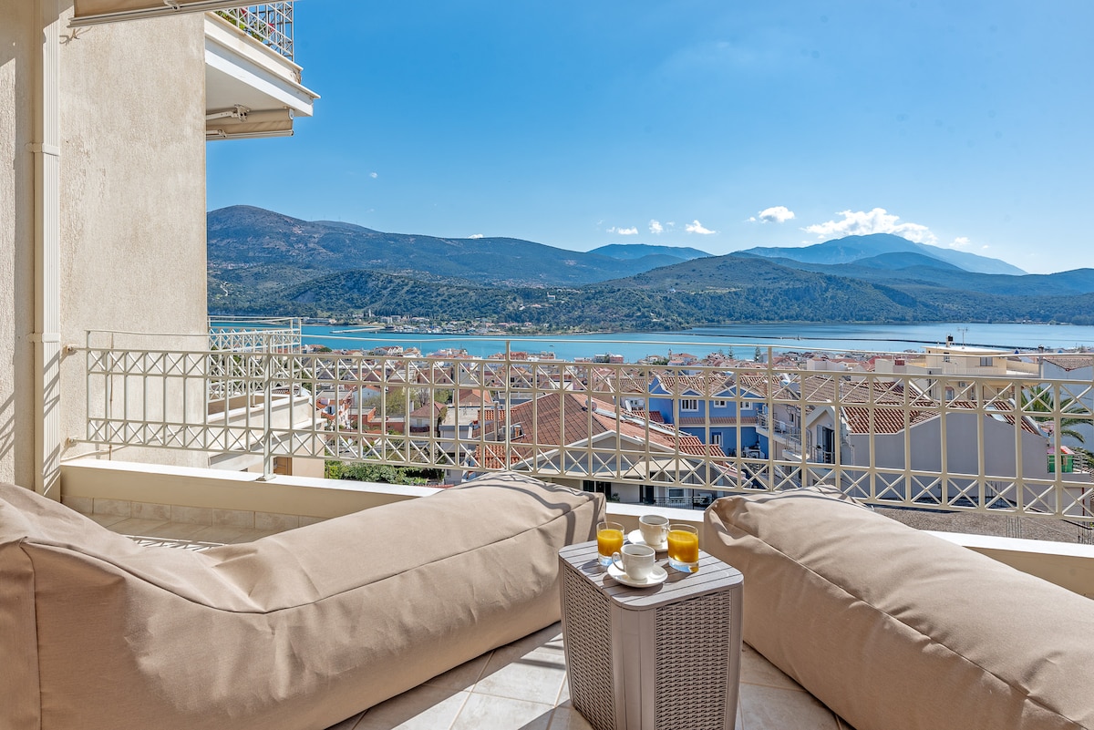 A balcony view showcases a stunning landscape of the Ionian Sea and surrounding mountains. Light-colored cushioned seating is arranged around a small table set with drinks, inviting guests to enjoy the serene atmosphere and picturesque scenery.
