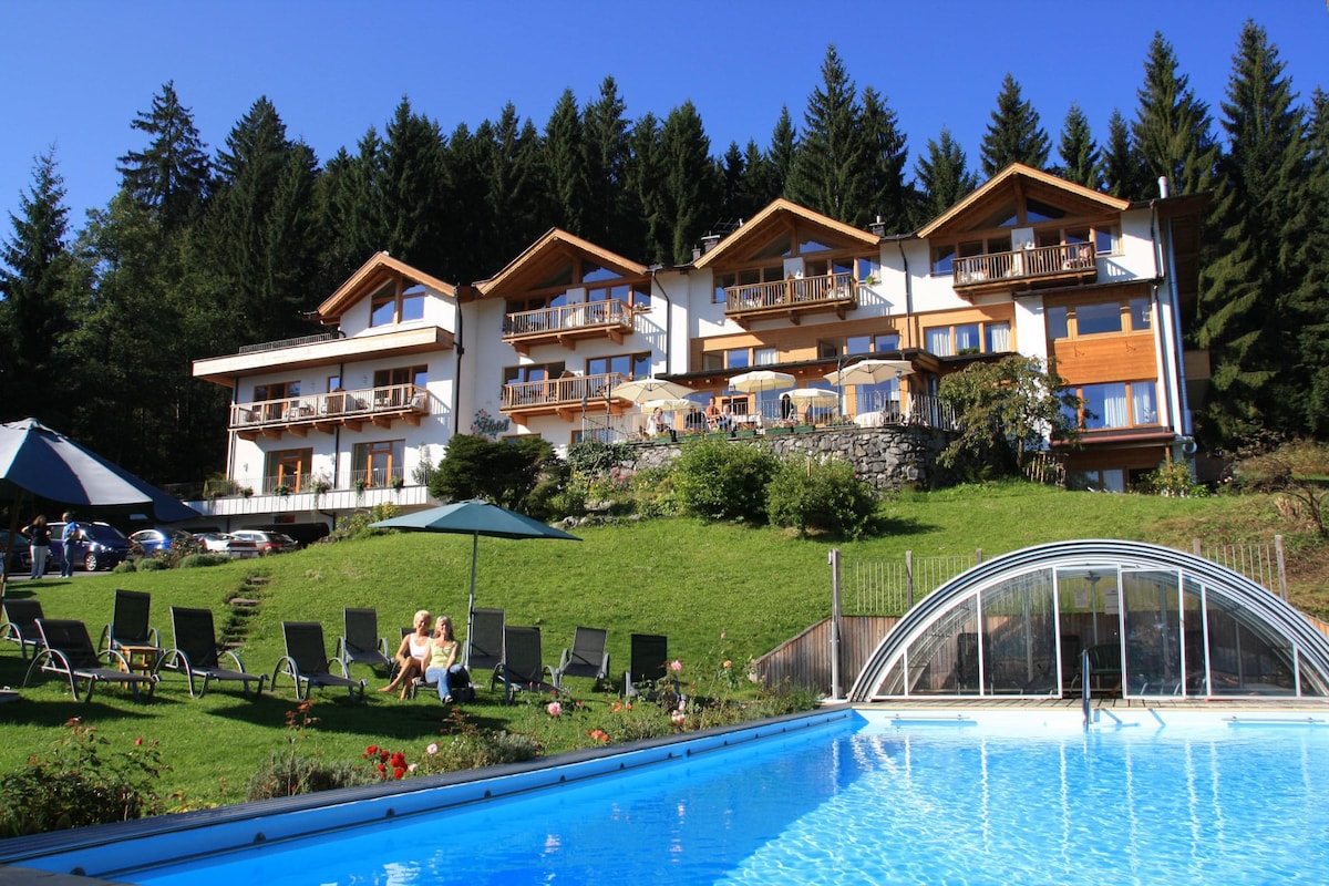 A garden hotel is set against a backdrop of lush trees, featuring a modern building with multiple balconies. Sun loungers are arranged on green grass, near a heated pool with a transparent retractable roof. The serene environment is complemented by the natural landscape.