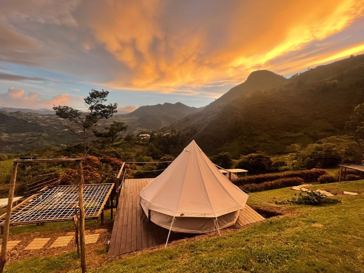 A spacious tent is set against a backdrop of rolling mountains and a vibrant sunset sky. The outdoor area features wooden decking and a grassy lawn, with scattered shrubs and plants enhancing the natural landscape.