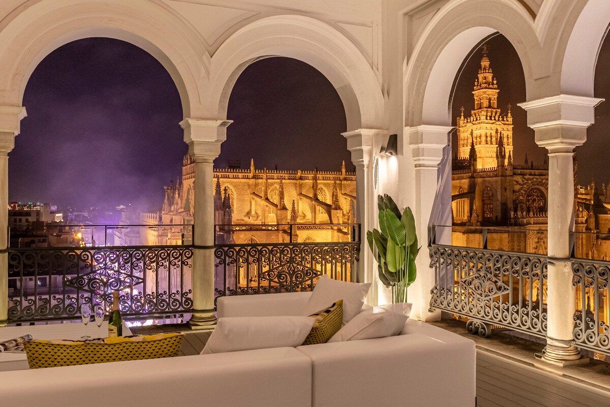 An elegantly furnished terrace is framed by arched columns, offering uninterrupted views of illuminated architectural landmarks at night. Soft seating options are arranged for comfort, complemented by greenery, creating a serene atmosphere for relaxation and contemplation of the city’s historic beauty.