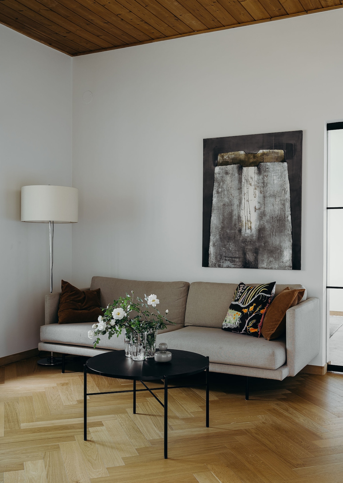 A light-filled living area features a comfortable beige sofa adorned with colorful cushions. A black coffee table sits in front, displaying a vase of fresh flowers. A large piece of wall art hangs above the sofa, complemented by a floor lamp beside it.
