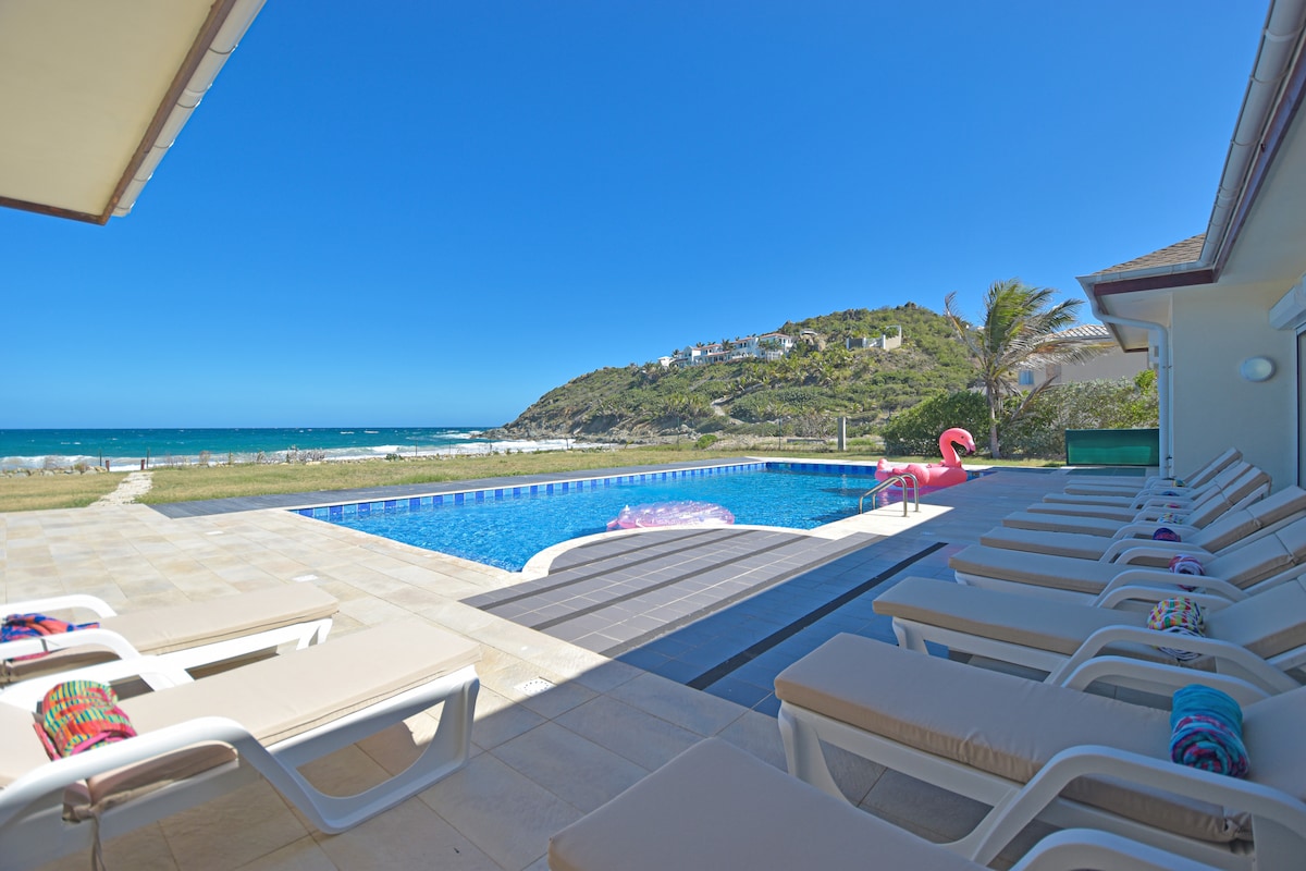 A spacious outdoor area features a private pool surrounded by lounge chairs. Bright towels are neatly arranged on the chairs. Green hillside and ocean views are visible beyond the pool, with a flamingo floatation toy resting nearby.