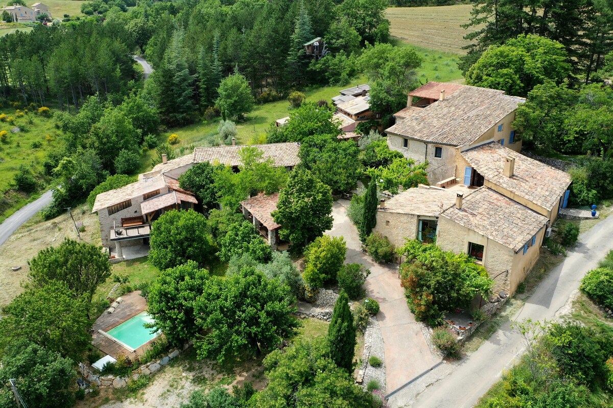 An aerial view of a rustic property surrounded by lush greenery. Several buildings with traditional stone facades and tiled roofs are nestled within the landscape, including a pool area. Trees and flowering plants provide a natural setting, enhancing the peaceful ambiance.
