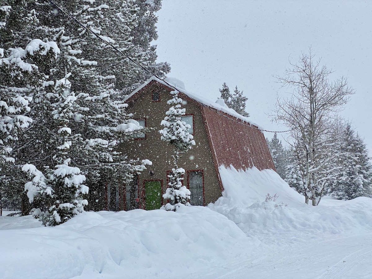 A charming cabin is surrounded by snow-covered trees, showcasing a sloped roof with a red metal finish. The ground is blanketed in fresh snow, and snowflakes fall gently, creating a serene winter atmosphere.