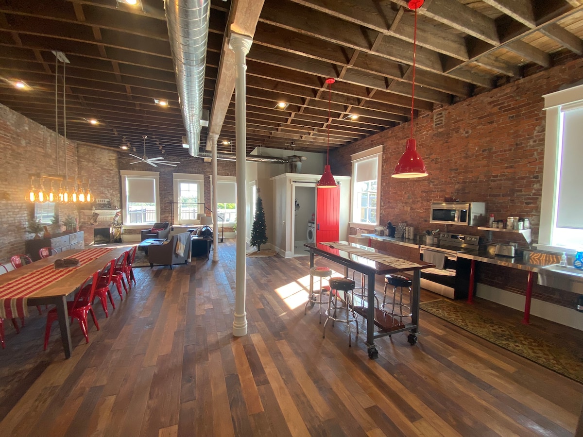 An open-concept great room features exposed brick walls and wood beam ceilings, with large windows allowing natural light. A rustic dining table surrounded by red chairs is positioned next to a modern kitchen area. A cozy seating arrangement with a sofa is visible in the background.
