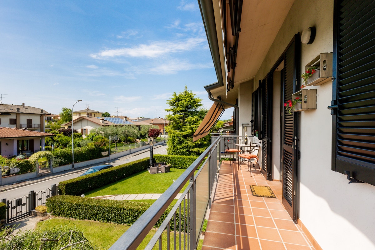 A balcony space is visible with a tiled floor and metal railing. Seating is arranged for two, and potted flowers are placed near the door. The surrounding area features well-kept gardens and residential buildings, with the street extending out towards greenery under a clear blue sky.