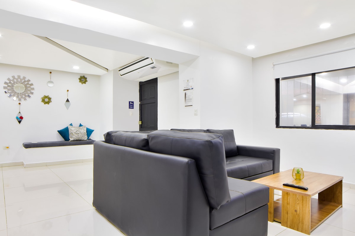 A modern living area features a large, dark sofa with sleek lines, positioned across from a wooden coffee table. Light-colored walls and glossy flooring enhance the space, while decorative wall hangings and cushions add subtle color. Natural light enters through a nearby window.