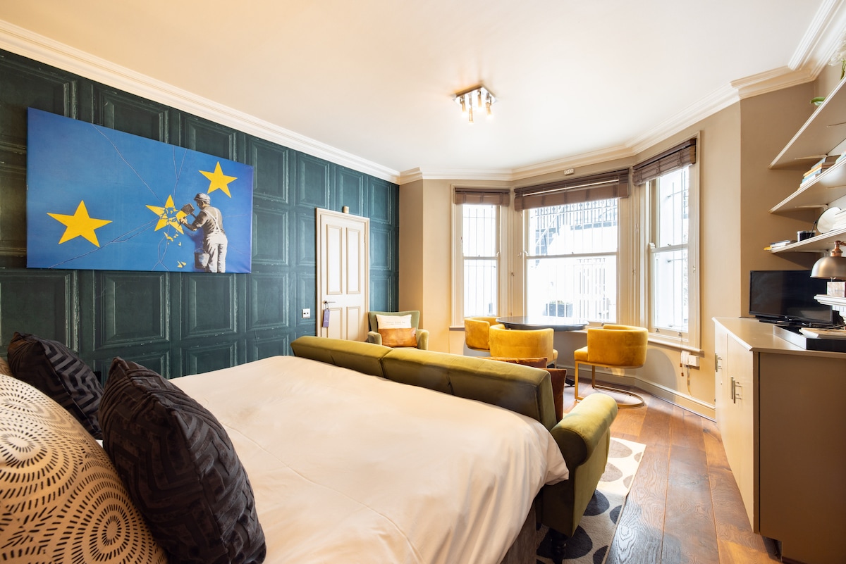 A studio space features a superking size bed with dark cushions, set against a backdrop of deep green paneling. A large art piece depicting blue and yellow stars adorns the wall. Two yellow chairs and a small table are positioned near large windows, allowing natural light to fill the room.