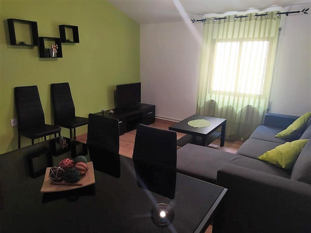A living area features a modern, dark table contrasting with light green walls. A plush gray sofa is positioned beside a bright window covered with sheer drapes, allowing natural light to filter in. Several black chairs are arranged around the table, creating a cozy seating arrangement.