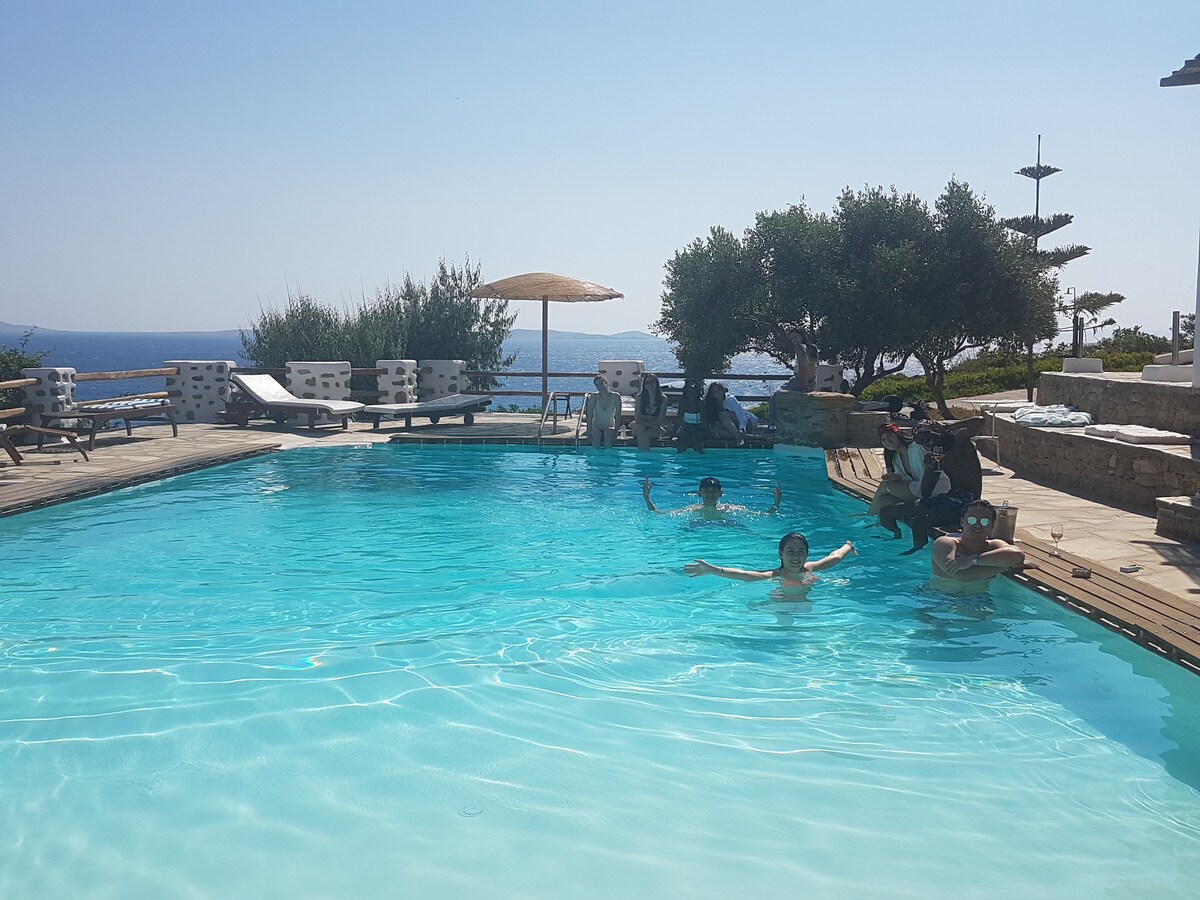 A serene swimming pool area features clear turquoise water surrounded by wooden decking. Sun loungers are arranged near the pool, with a large umbrella providing shade. Gentle hills and the sea are visible in the background, creating a tranquil environment.