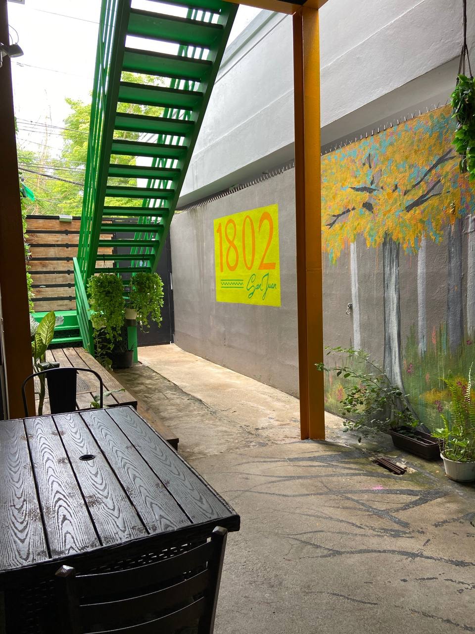 A vibrant outdoor corridor features a striking green staircase and a colorful mural depicting trees. Potted plants are positioned along the wall, adding a touch of nature to the space. A rustic wooden table and chairs are visible, offering a spot to relax.
