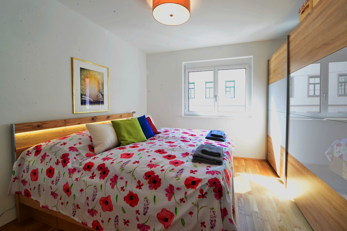 A bright bedroom is furnished with a spacious bed adorned in floral bedding. Two windows allow natural light to fill the room. A large wardrobe reflects the décor, and decorative pillows in various colors enhance the inviting atmosphere.