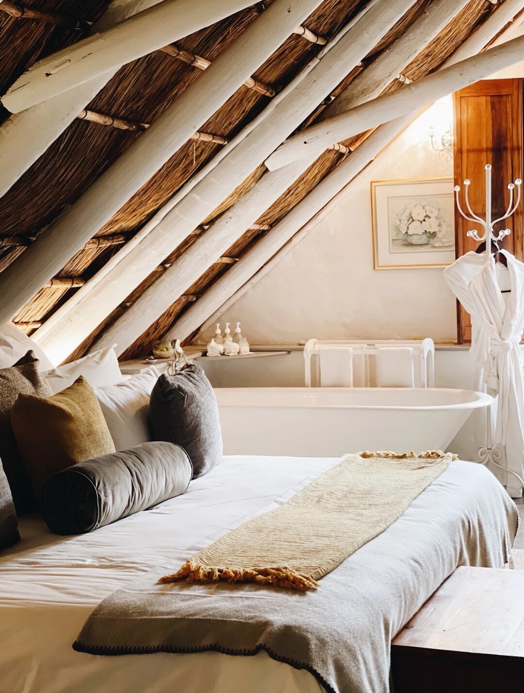 A cozy bedroom features a spacious bed adorned with decorative pillows. Exposed wooden beams create an inviting atmosphere. A freestanding bathtub is positioned nearby, enhancing the room's rustic charm. Soft natural light illuminates the space, highlighting the decor's neutral tones.