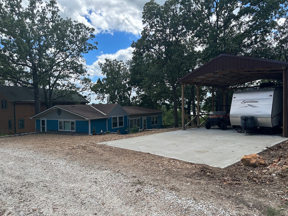 The image captures a spacious property with a vibrant blue house adjacent to a covered carport. The newly paved concrete pad is visible beneath the carport, providing ample parking space. Surrounding trees offer a natural setting, and a gravel area leads towards the house.