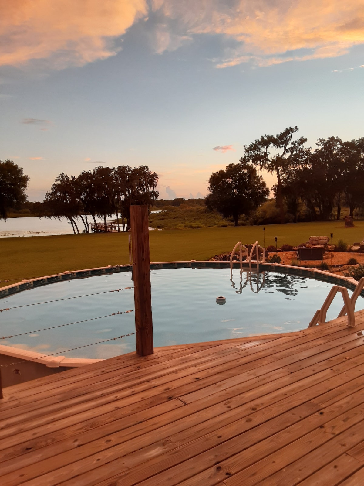 A serene view of a circular swimming pool is presented, surrounded by a wooden deck. The calm water reflects the sky, while lush greenery and a water body are visible beyond the pool area. A gentle sunset casts soft colors over the scene.