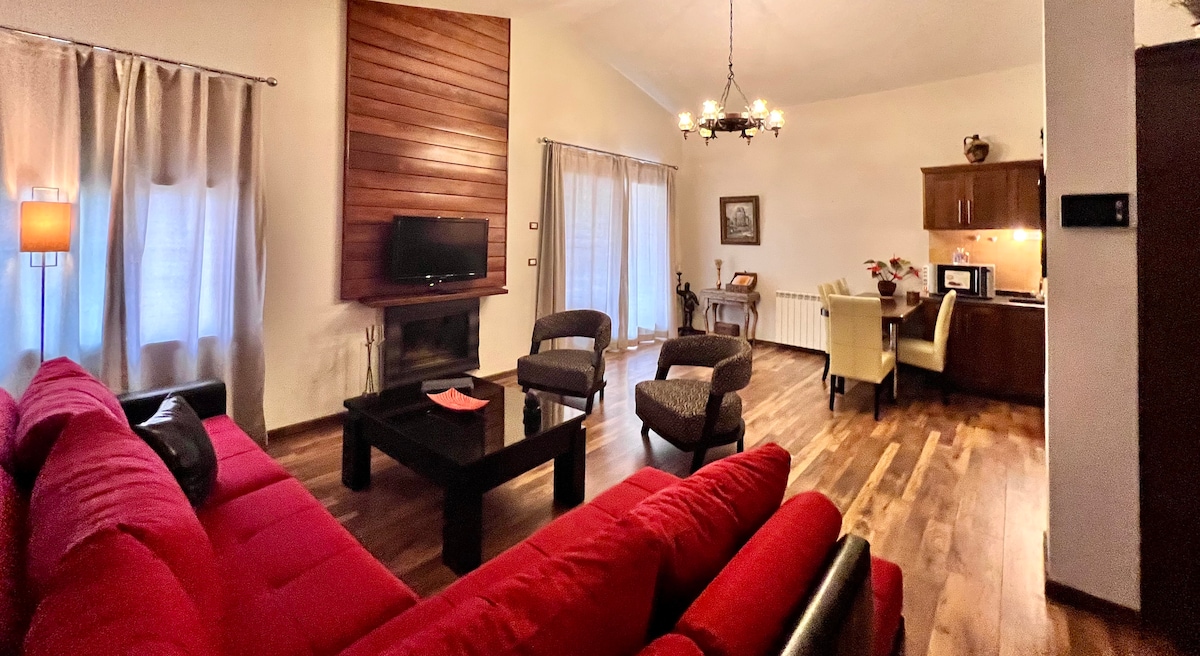 A spacious living area is enhanced by warm wood flooring and soft, natural light filtering through sheer curtains. A red sofa and two comfortable chairs are arranged around a sleek black coffee table, with a subtle fireplace and dining area visible in the background.