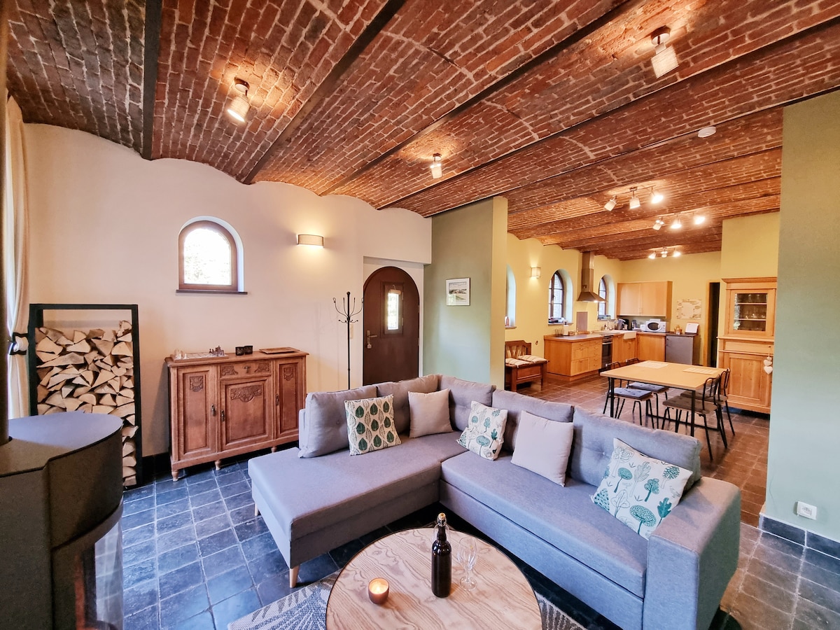 A spacious living area features a large, gray sectional sofa adorned with decorative cushions. Exposed wooden beams and a brick ceiling create a warm ambiance. A coffee table is set in front, while a dining area and kitchen are visible in the background, showcasing wooden cabinetry.