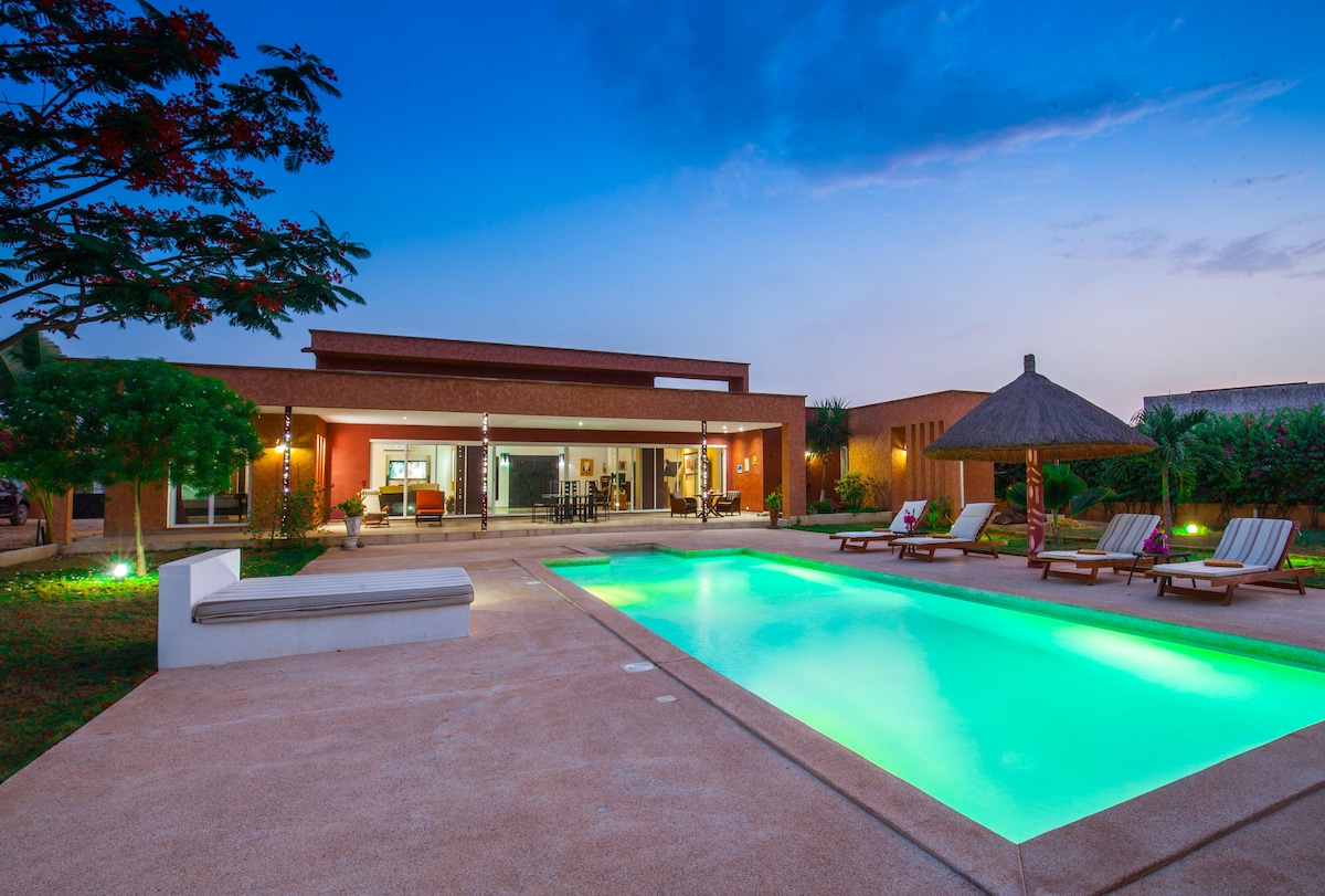 A modern villa is depicted with a spacious swimming pool surrounded by relaxation chairs. The structure features large windows inviting natural light. A thatched roof provides shade, and landscaped greenery enhances the outdoor setting, creating an inviting space for relaxation.