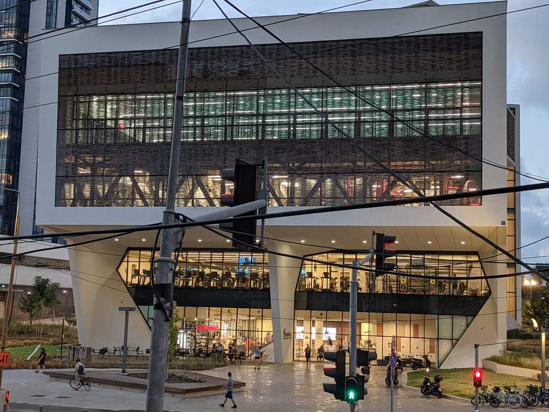 The modern building features a striking geometric design with large glass windows revealing an interior space filled with activity. The structure is supported by sleek, angular columns, with illuminated signage visible at ground level. Pedestrians and cyclists can be seen in the surrounding area.
