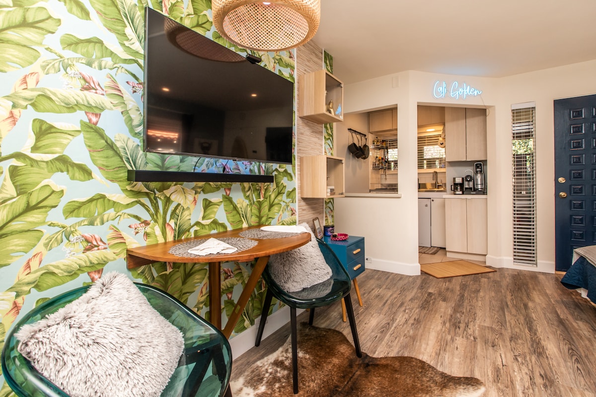 A cozy living area features a large television mounted on a tropical leaf-patterned wall. A wooden table is paired with two green chairs, while soft textures are brought in with a fur rug. The kitchen area is visible, with modern appliances and cabinetry.