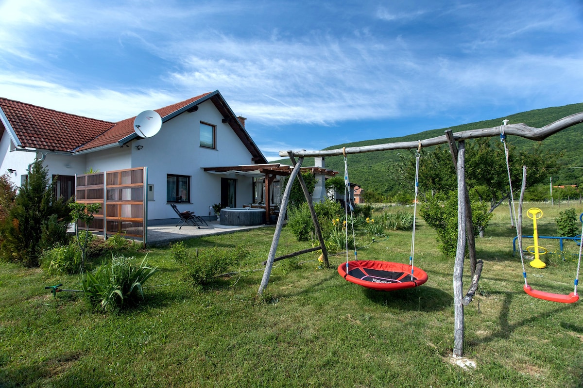 A charming holiday home is surrounded by a well-maintained garden, featuring green grass and blooming plants. A swing set with a round red swing and a yellow swing is positioned nearby, enhancing the outdoor space. The house showcases a sloped red roof and a patio area.