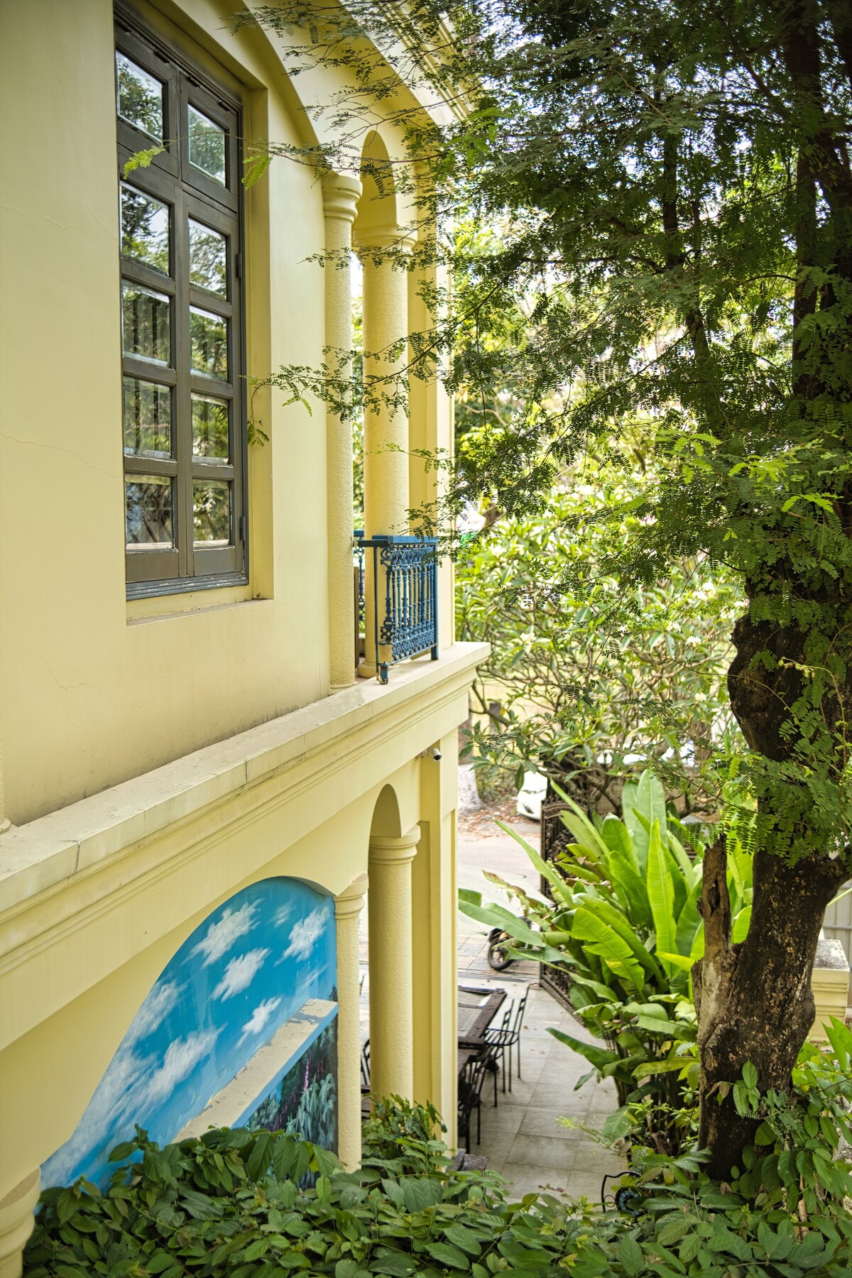 A glimpse of the villa's exterior reveals a charming yellow facade adorned with large arched windows and a blue balcony railing. Lush greenery and tall trees frame the scene, contributing to the villa's serene atmosphere. A hint of outdoor seating can be seen in the background.