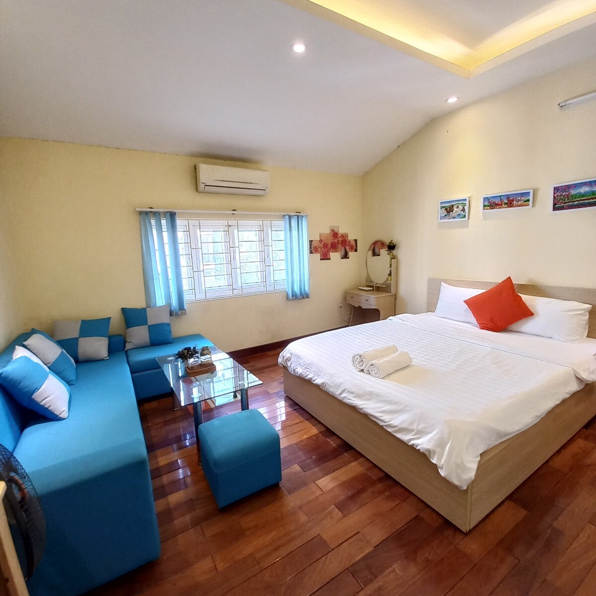 A well-lit room features a spacious bed with a white duvet and an orange accent pillow. A comfortable seating area with blue cushions is positioned near a glass table. Two windows allow natural light, while air conditioning and a dressing table add convenience.