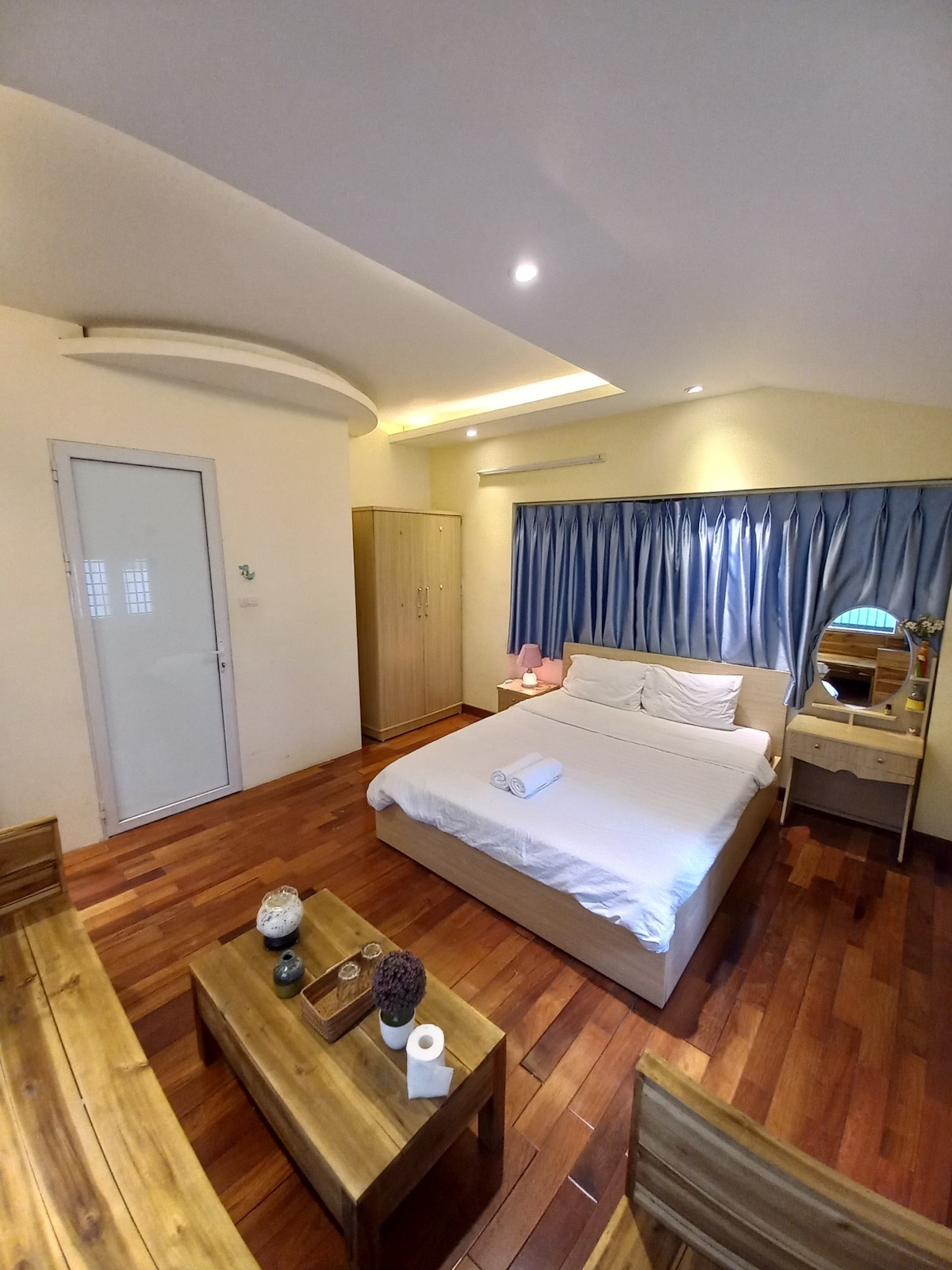 A cozy room features a spacious bed with fresh linens, complemented by wooden accents on the floors and furniture. A large window with blue drapes allows natural light to fill the space. A wardrobe and a small vanity with a mirror are also visible.