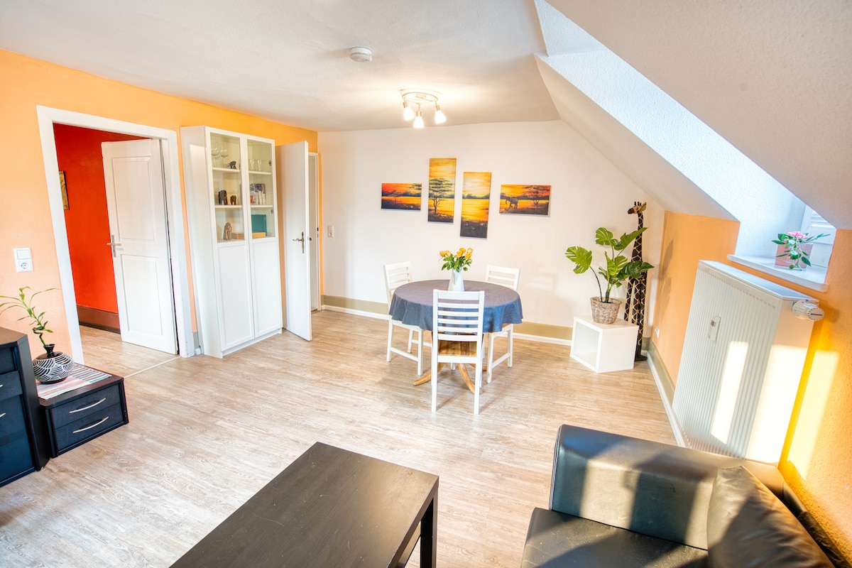 The living space features a dining area with a round table and four white chairs, complemented by framed artwork on the orange wall. A black sofa sits nearby, while natural light streams in from a window. A potted plant adds a touch of greenery.