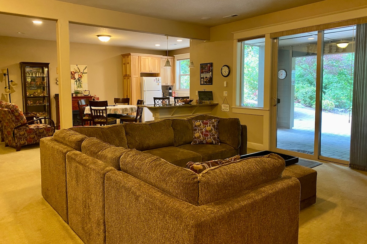 A spacious living area features a large sectional sofa with patterned cushions, offering a comfortable place to relax. Natural light enters through sliding glass doors that lead to the outdoor space, while an adjacent dining area is visible, along with the kitchen in the background.